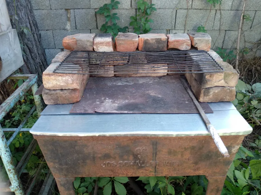 Postavený venkovní gril ze starých cihel s kovovým roštem nahoře, umístěný na stinném místě u zídky. Gril stojí na kovové konstrukci s plechem a kolem něj rostou zelené rostliny.