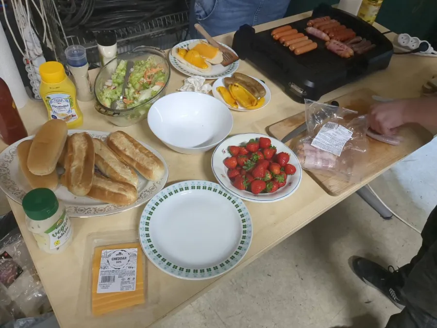 Na stole je připravena jídla k neformálnímu stolování. Jsou zde housky, salát, jahody a plátky sýra. Na deskovém grilu se opékají párky. Vedle grilu se krájí další ingredience. Na stole jsou také láhve s kečupem a hořčicí.