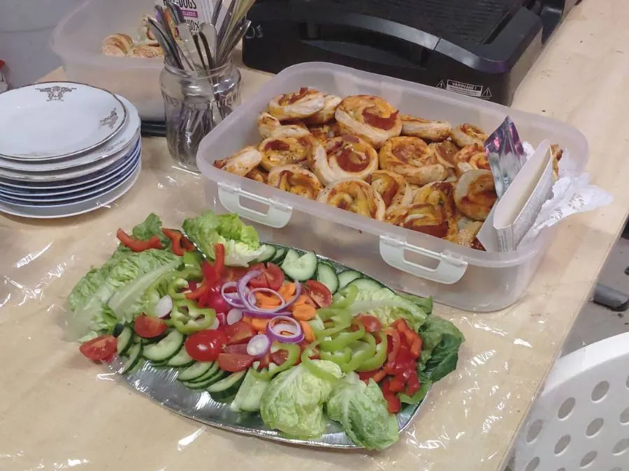 Stůl s podnosem plným čerstvé zeleniny včetně okurek, paprik, rajčat a salátu. Vedle se nachází krabice s veganskými pizza šneky. V pozadí je vidět několik naskládaných talířů a sklenice s příbory.
