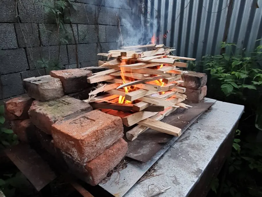 Dřevěná hranice připravená k zapálení na venkovním grilu z cihlového a kovového podkladu. Plameny a kouř stoupají nahoru, zatímco v pozadí je vidět zeď z betonových bloků a zelené rostliny.