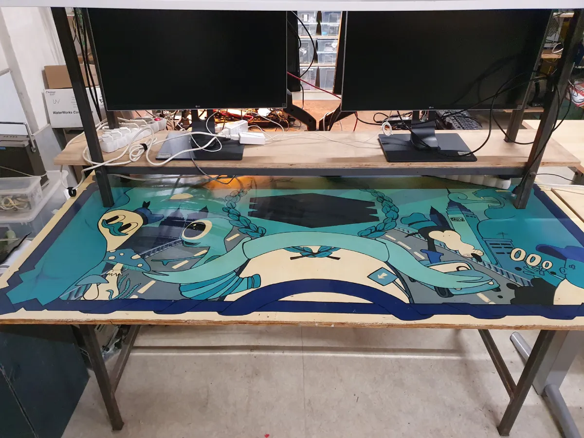 Deep lore, core memories. Stůl s barevně ilustrovanou deskou zobrazující abstraktní motiv v odstínech modré a béžové. Na stole jsou dva monitory a zamotané kabely.