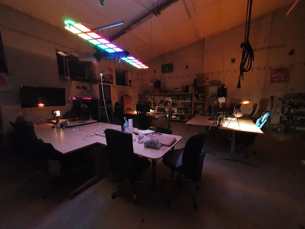 Hackerspace Base48 ve večerní atmosféře s několika pracovními stoly a počítačovými židlemi. Nad stoly visí satelis sloužící jako LED světlo. V pozadí jsou vidět police s vybavením a nástroji.