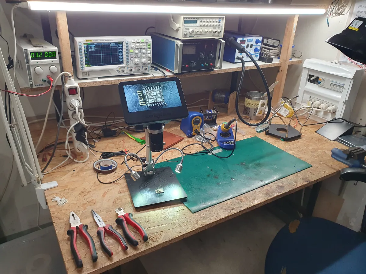 Pracovní stůl v hackerspace s různými elektronickými zařízeními, včetně osciloskopu, napájecího zdroje a pájky. Na stole jsou také kleště a mikroskop s displejem zobrazujícím detailní pohled na mikrochip ESP32. Stůl je dobře osvětlen zářivkou nad ním.