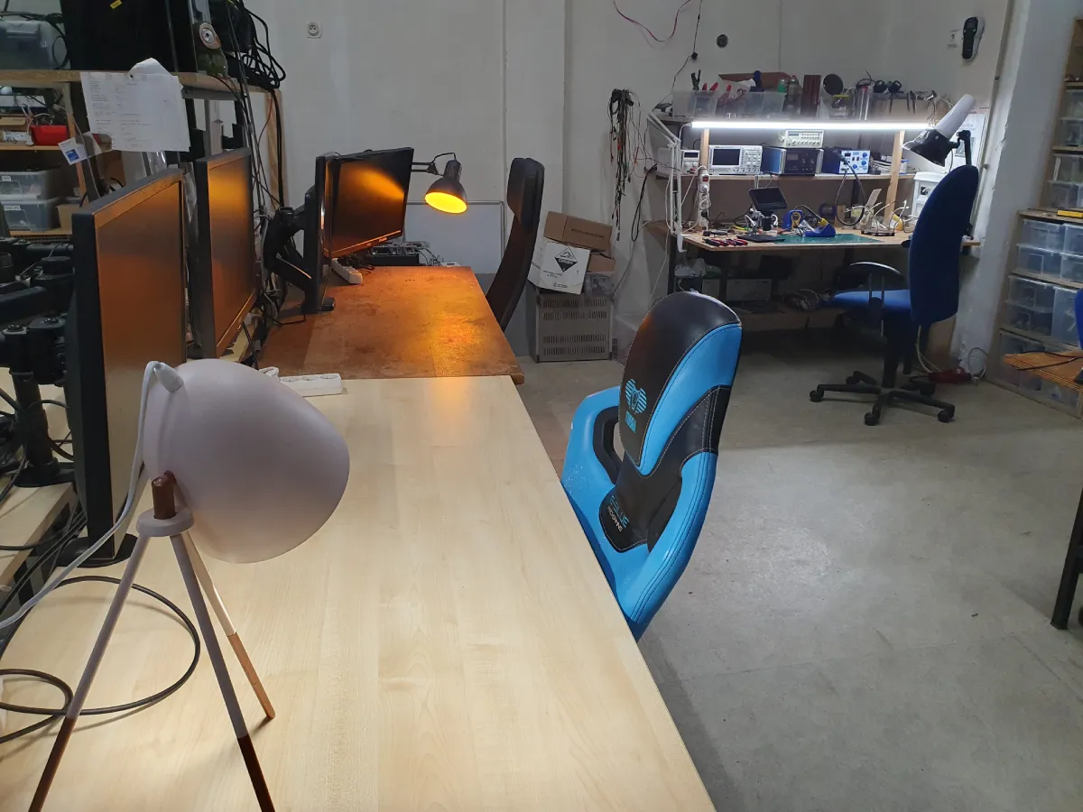 Pracovní prostor s několika stoly vybavenými počítači a monitory, židlemi a lampami. V pozadí je vidět další pracovní stůl s elektronikou, nástroji a měřicími přístroji.